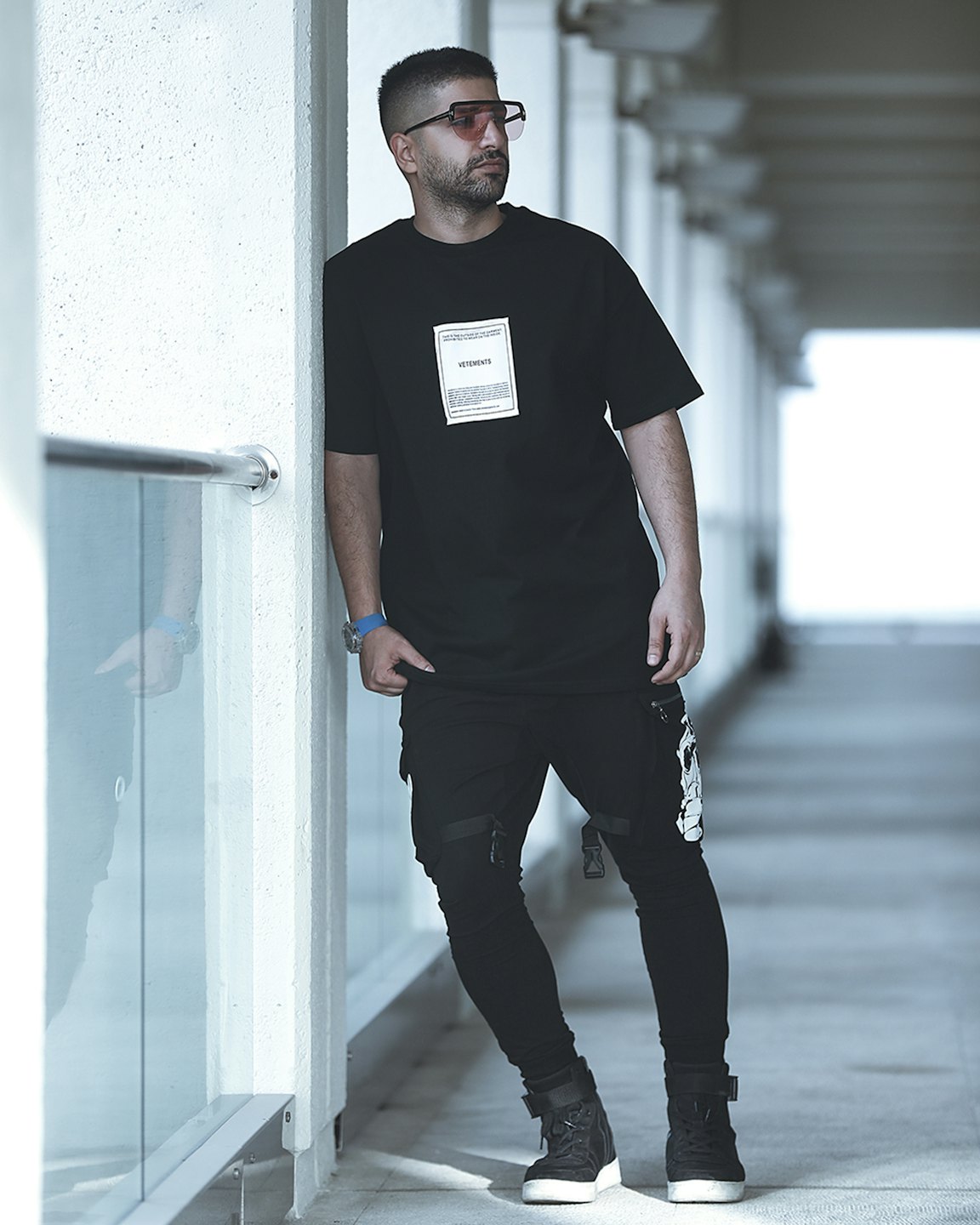 Man in black streetwear leaning against a white concrete wall.