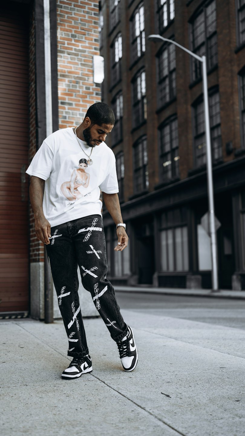 Young Black man on city sidewalk wearing graphic tee and patterned jeans.