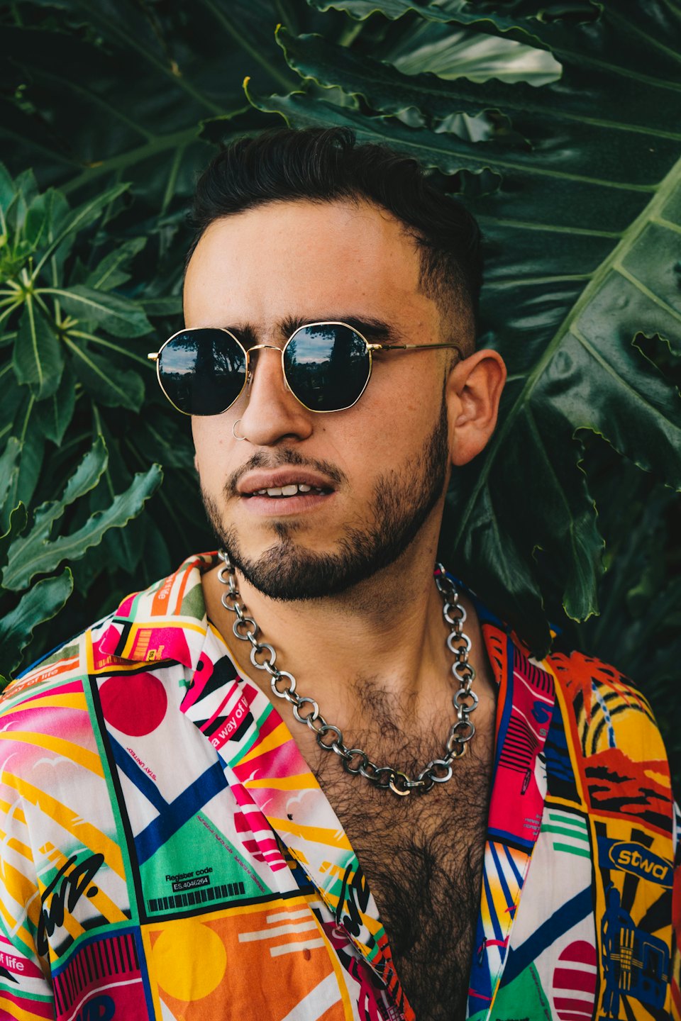 Man wearing round sunglasses and a patterned shirt stands outdoors.
